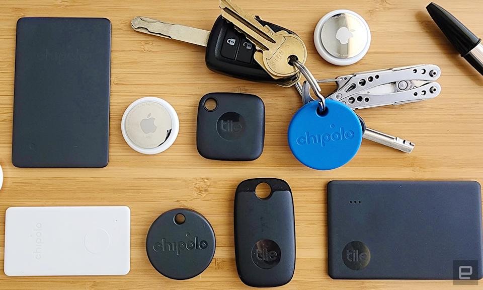Een assortiment bluetooth-trackers gerangschikt in een rooster op een houten achtergrond. Trackers zijn onder andere zwarte Tile-trackers in verschillende vormen, twee zilveren en witte AirTag-trackers en een ronde blauwe Chipolo-tracker bevestigd aan een sleutelbos met een multitool-sleutelhanger.