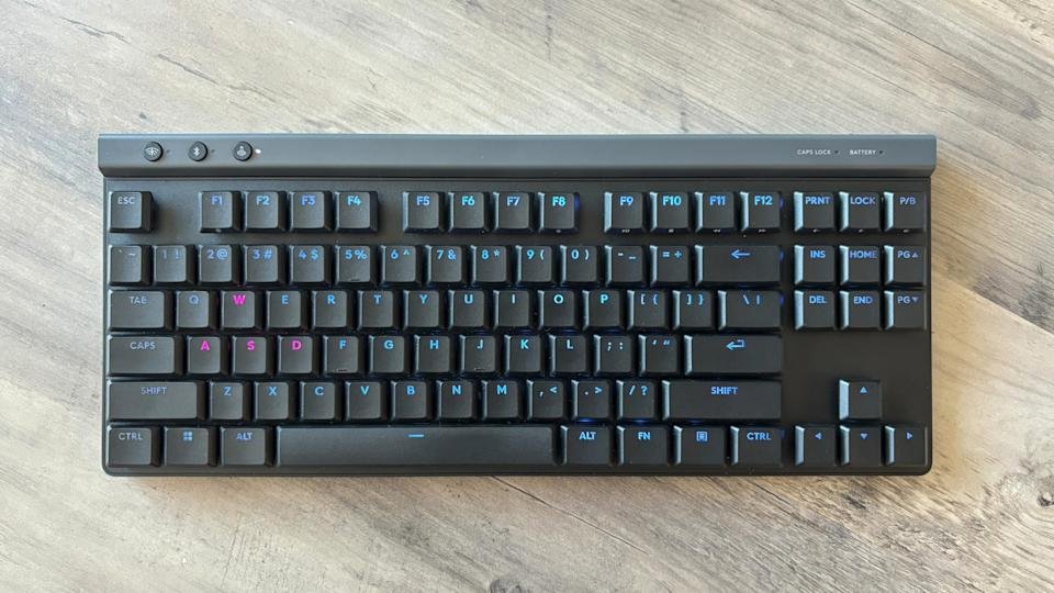 Een zwart gamingtoetsenbord met lichtblauwe RGB-verlichting, de Logitech G515 TKL Lightspeed, ligt op een bruine houten tafel.