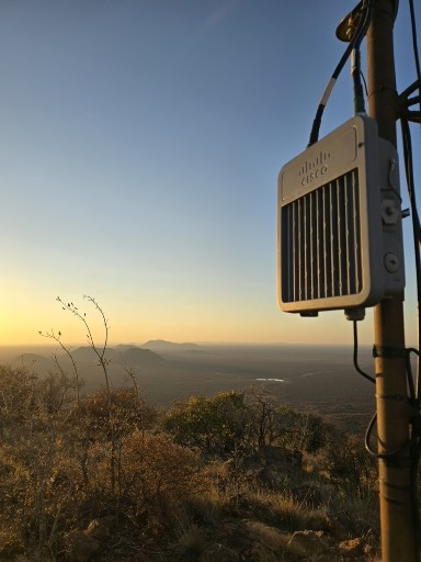 Een buitennetwerk van outdoor netwerkapparaat ligt prominent op een heuvel, met uitzicht op een enorm panoramisch landschap ondergedompeld in het warme gouden licht van zonsopgang of zonsondergang.