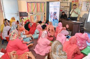 Een man spreekt in een microfoon tegen een grote groep vrouwen die op de grond zitten, met een "Cyber Suraksha Plus" spandoek op de achtergrond.