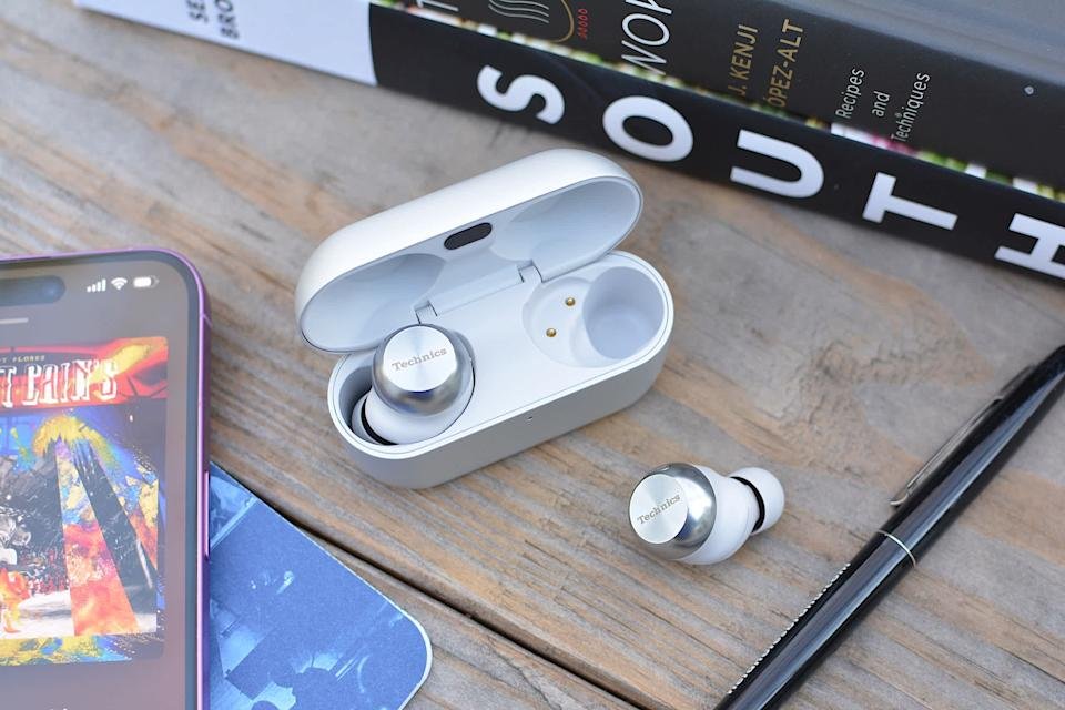 Een paar Technics AZ100-oortelefoons staan ​​op een houten bureau, naast een iPhone en een paar boeken.