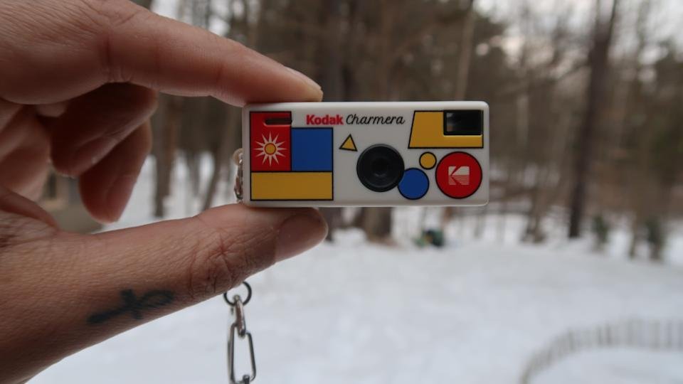 Er wordt een Kodak Charmera tussen twee vingers getoond, met sneeuw en bomen op de achtergrond