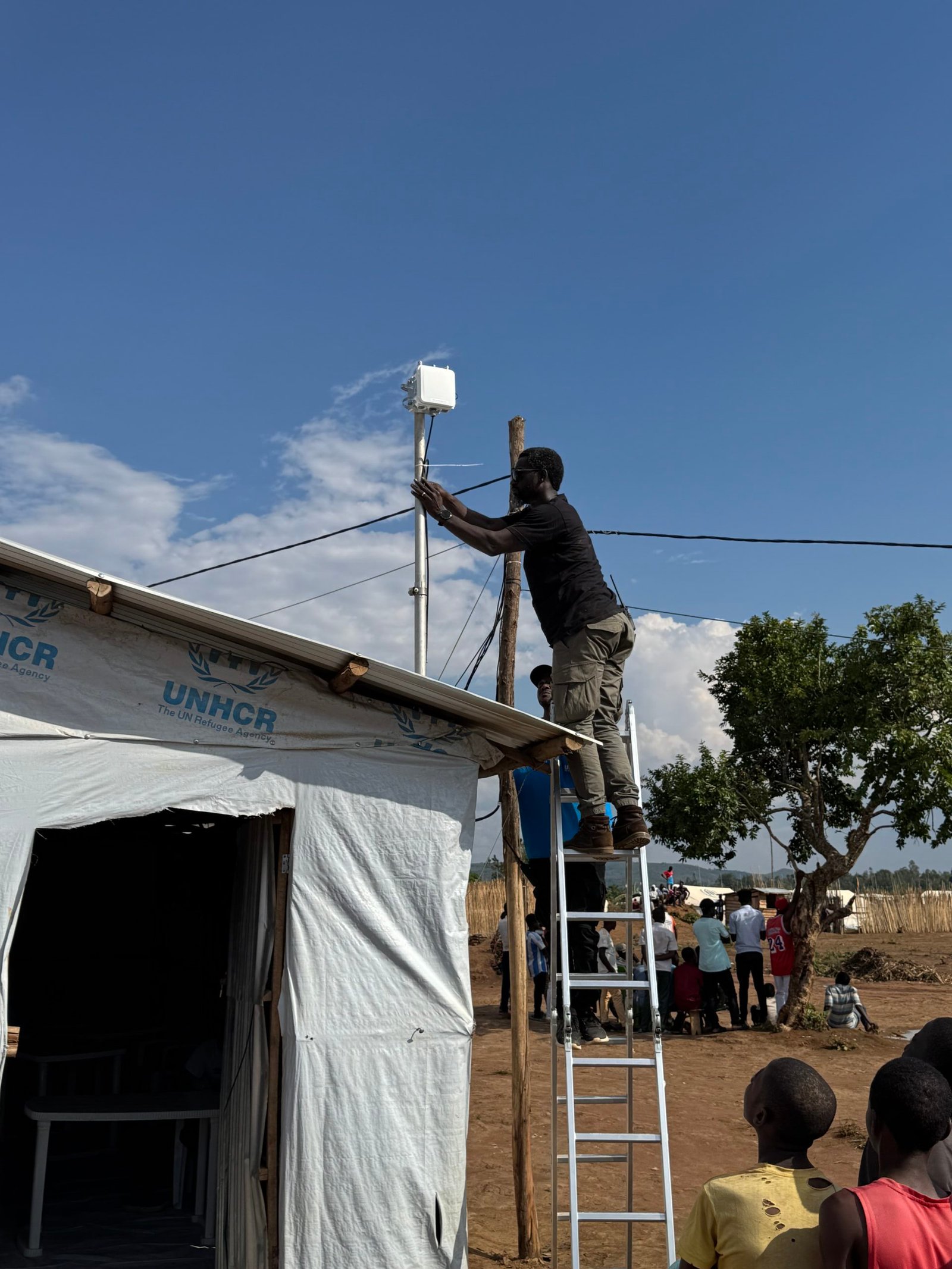 Een man staat op een ladder om een ​​antenne te installeren boven een UNHCR-tent op een vluchtelingenkamp in Burundi. Verschillende mensen kijken naar hem om goed te kijken, terwijl anderen op de achtergrond onder een boom zitten.