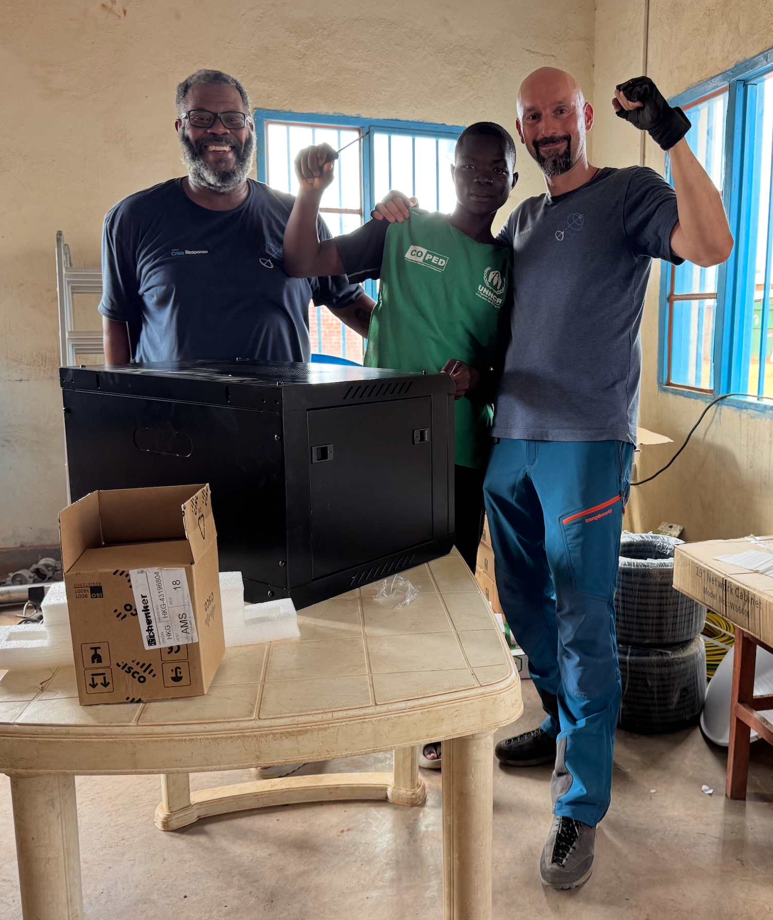 Drie mannen staan ​​voor technologische apparatuur in een kantoor in het vluchtelingenkamp Musenyi in Burundi. Alle drie de mannen glimlachen en de mannen in het midden en rechts heffen hun vuisten op in besef.