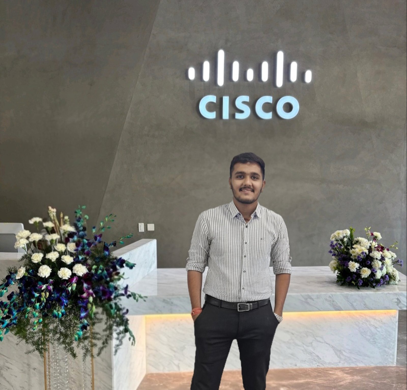 Een man glimlacht in de lobby van een modern kantoor onder een helder Cisco-logo, met aan weerszijden marmeren toonbanken en bloemstukken.
