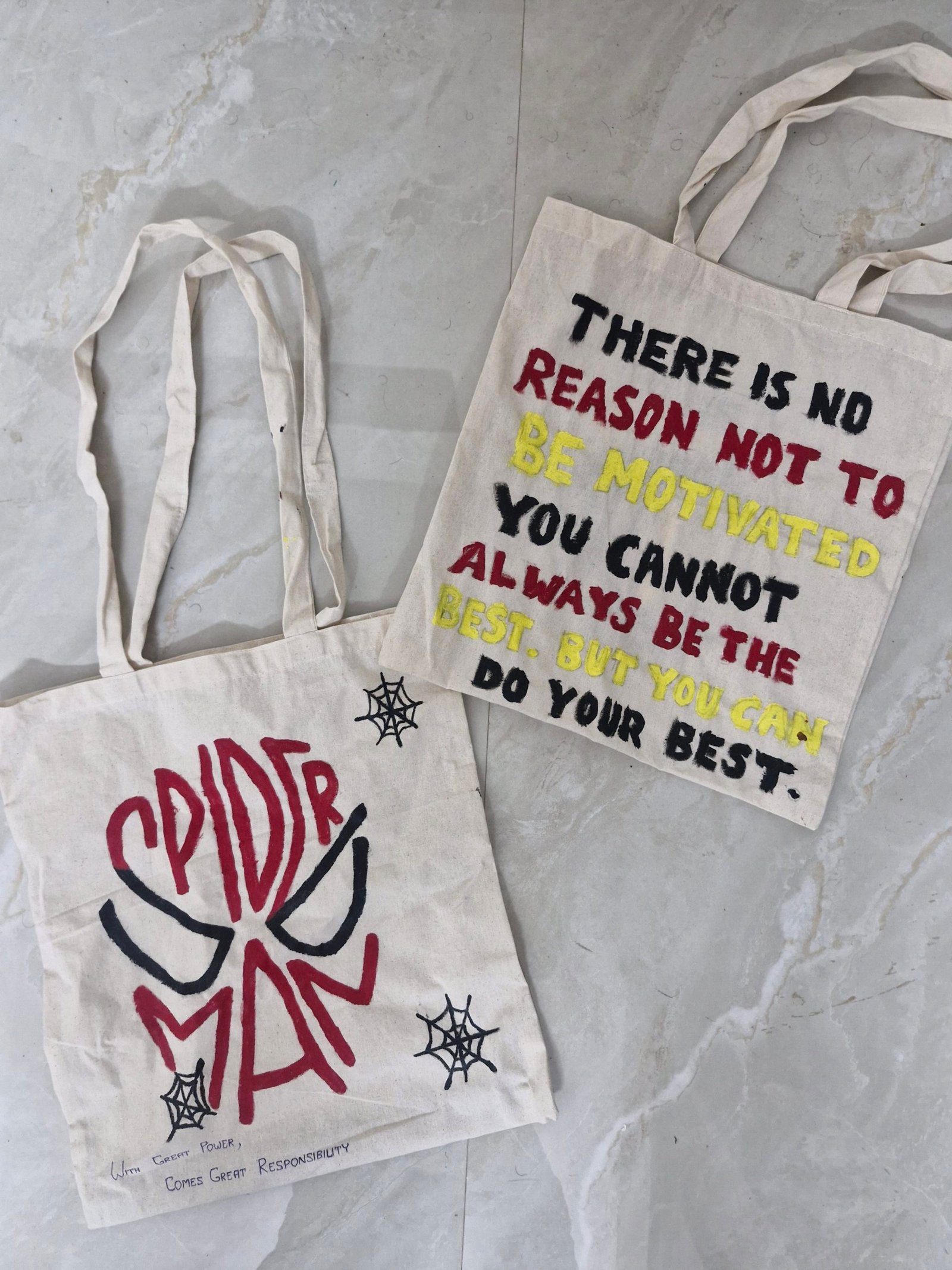 Twee canvas tassen op een tegelvloer: één met een tekening van Spider-Man en de andere met een motiverende quote geschreven in zwart, rood en geel.