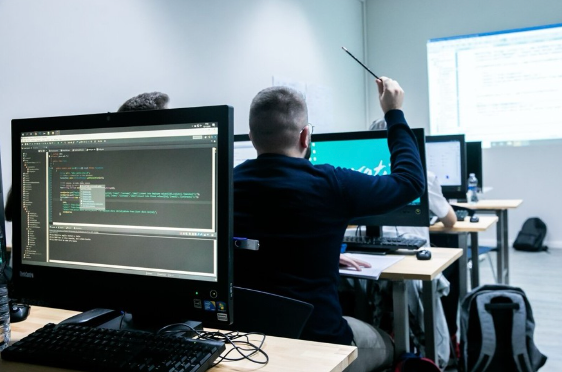 Deze afbeelding is van een klaslokaal in computerlokaalstijl. Links staat een groot computerscherm. In het midden steekt een man (van achteren) die achter een computer zit zijn hand op.
