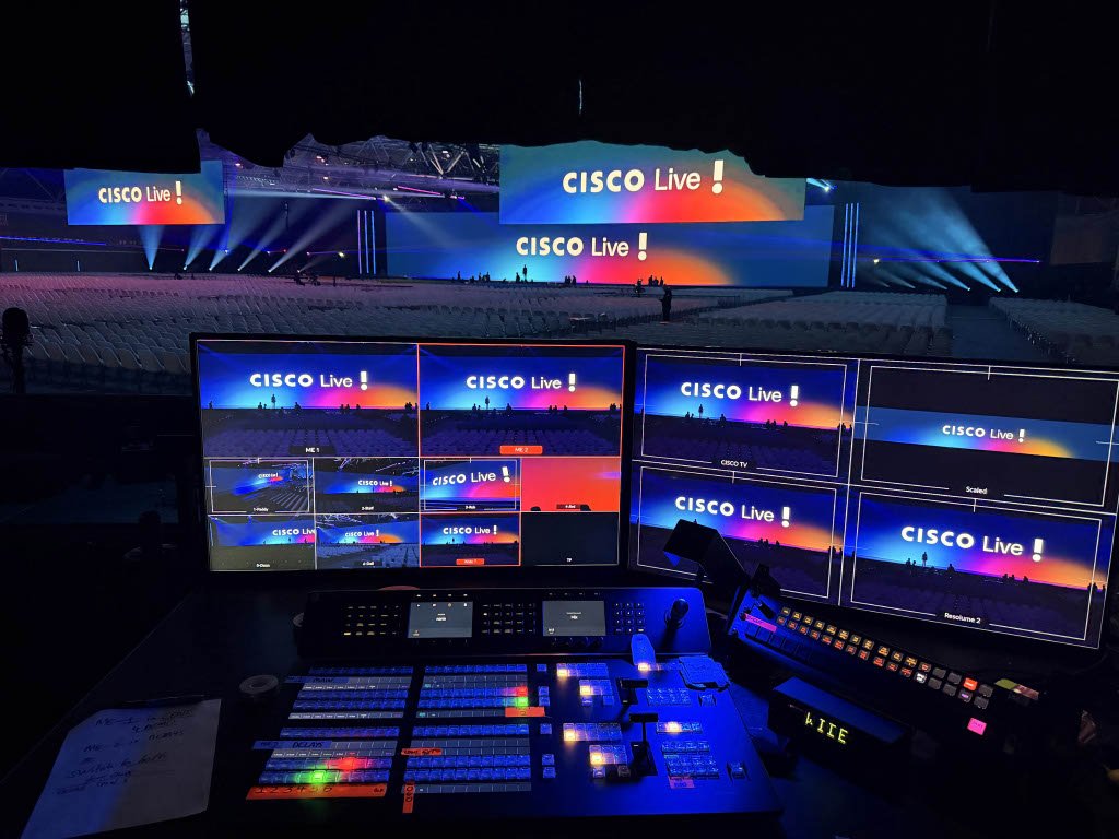 Uitzicht op de productiecontrolekamer van een groot evenement, met meerdere monitoren die het podium tonen en een mengpaneel op de voorgrond, met uitzicht op een goed verlicht podium en lege zitplaatsen voor het publiek.