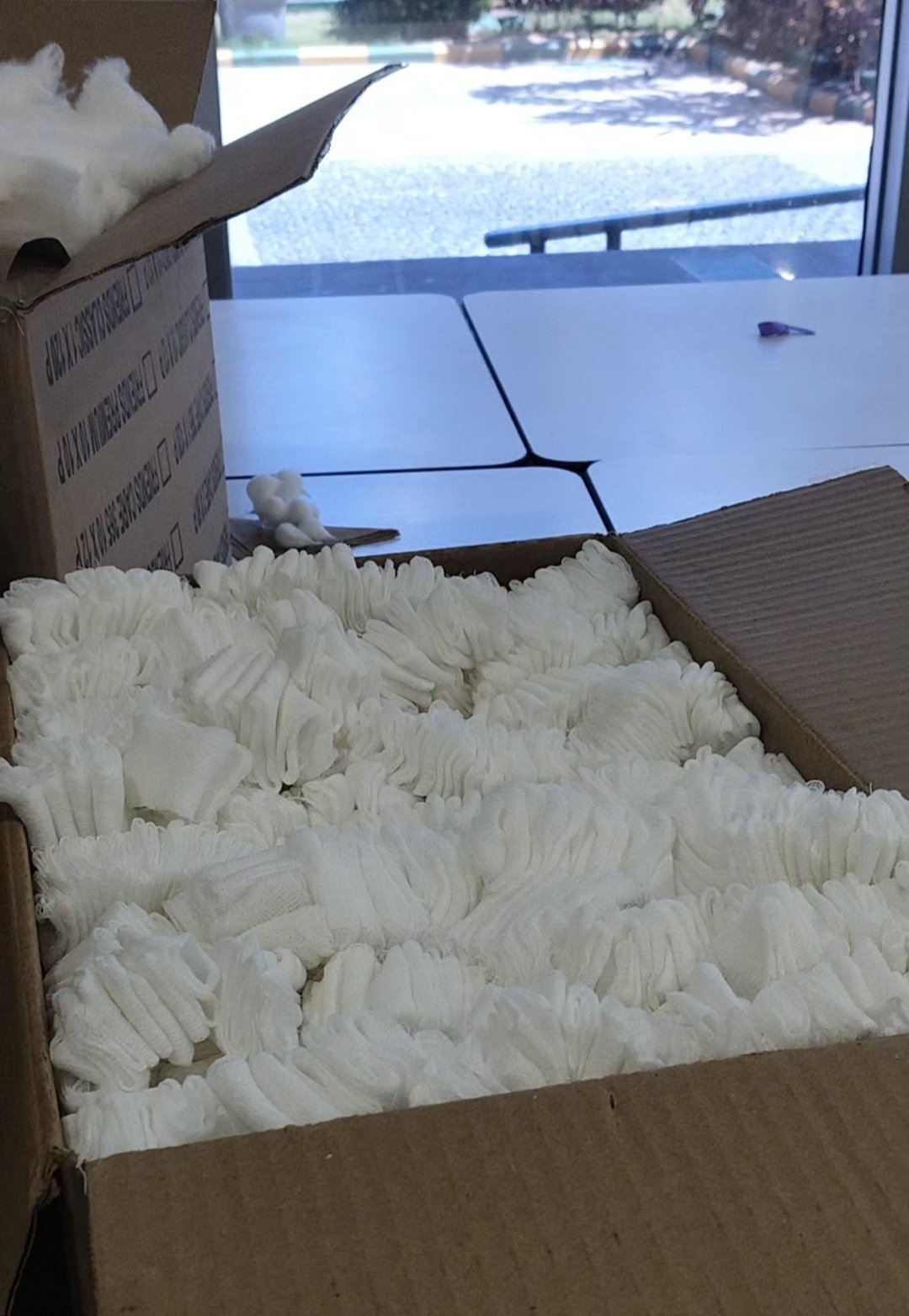 Open kartonnen dozen vol spullen of witte stoffen handschoenen, keurig opgestapeld op een tafel bij een raam, met verpakkingsmateriaal zichtbaar op de achtergrond.