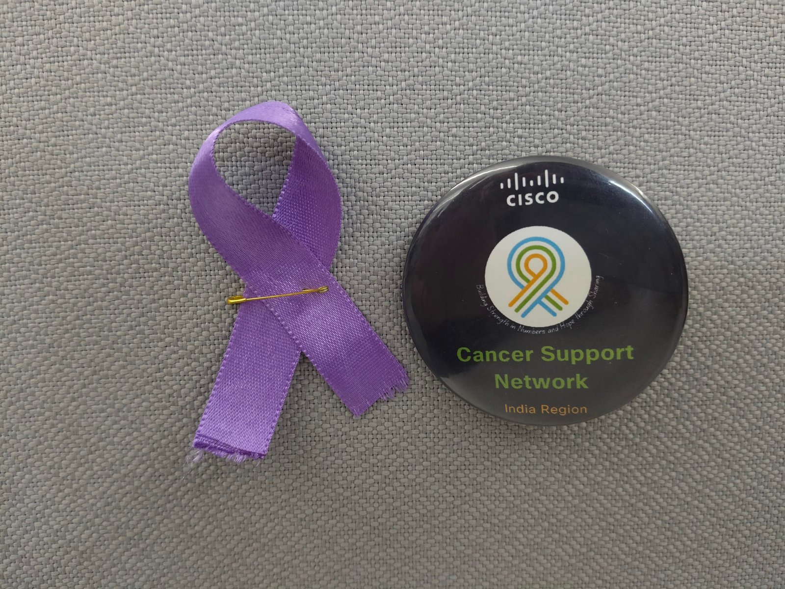 Een paars bewustzijnslint vastgemaakt naast een gelabelde Cisco Cancer Support Network-knop 'India Region', geplaatst op een achtergrond van grijs textiel.